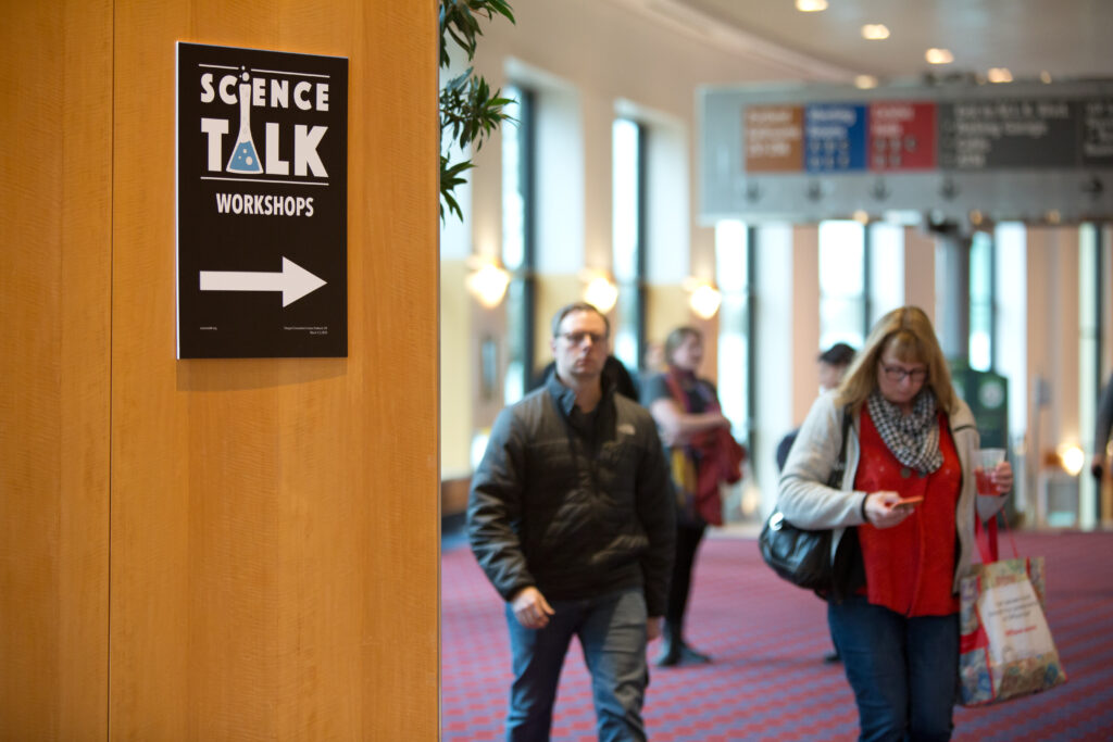 Door opening into room where people are gathering. Sign on the door has the words "Science Talk" and an arrow pointing into the room and the people within,