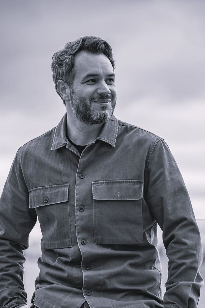 A black and white photo of a smiling man with short hair and a short beard in a button up shirt