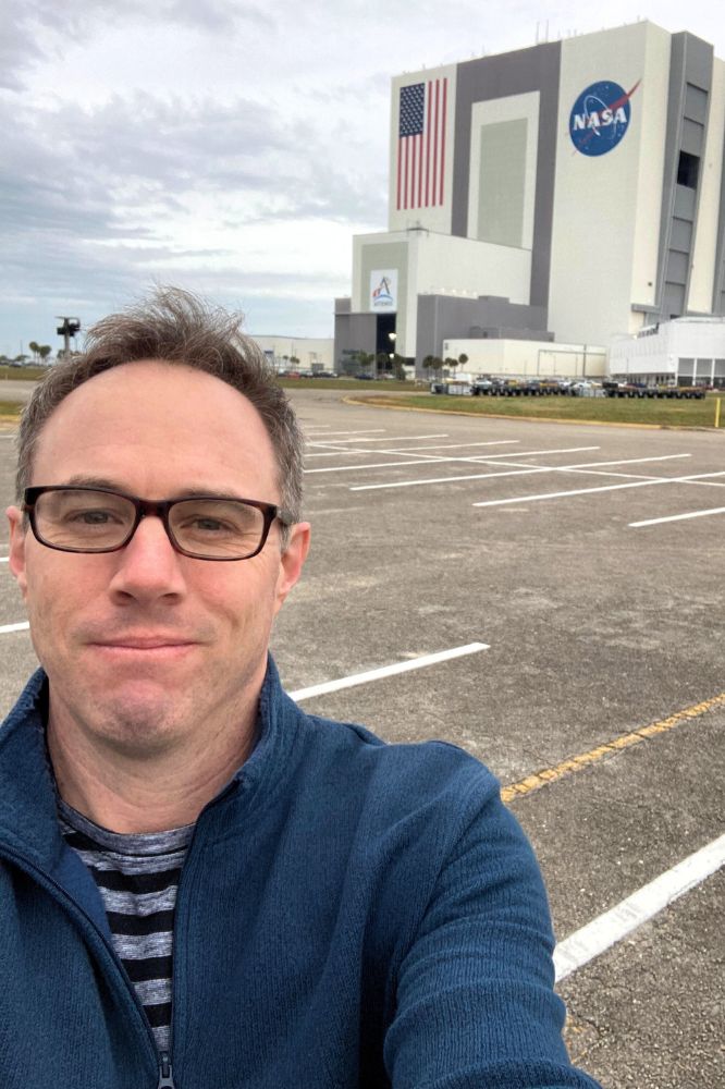 A man in glasses stands in a parking lot in front a large building with a big NASA logo and American flag on it