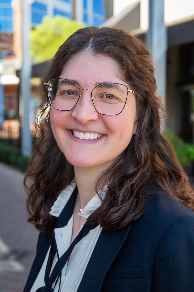 Photo of a smiling woman wearing glasses and a blazer and standing in front of a building