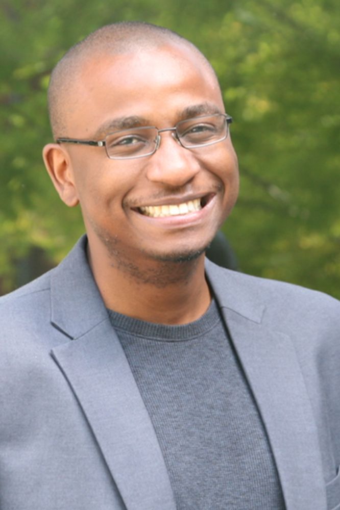 A photo of a smiling man with glasses, a blazer and a grey t-shirt