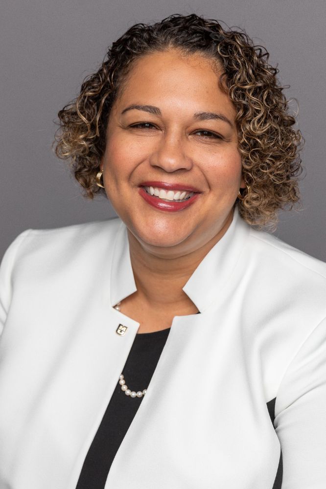 A professional headshot of a woman with curly hair in a white blazer and black shirt